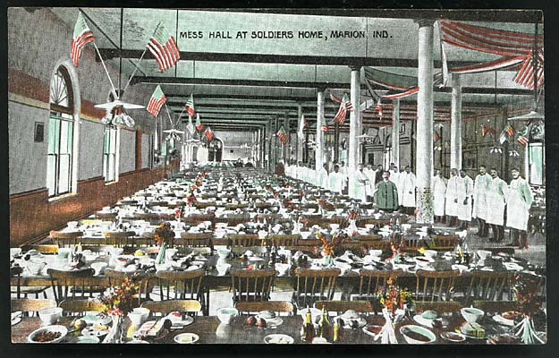 Mess hall at the Soldiers\' Home. Waiters, in white, are lined up along side of room, tables are set and prepared for a meal, flags and red & white bunting decorate the room. ca. 1910.